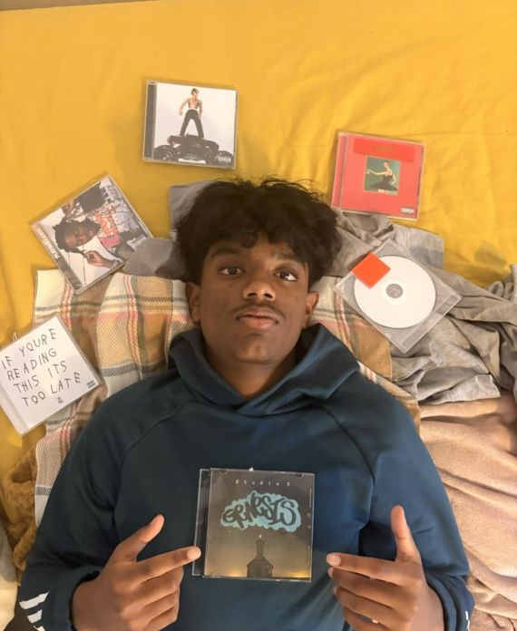 Aaron Cool lying on a bed of CD covers. He points at a CD cover on his chest which has "genesis" written on it