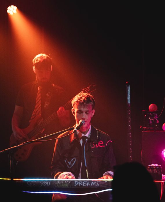 Keane Baldwin singing into the microphone while playing the keyboard in orange-red lighting. A guitarist plays behind him.
