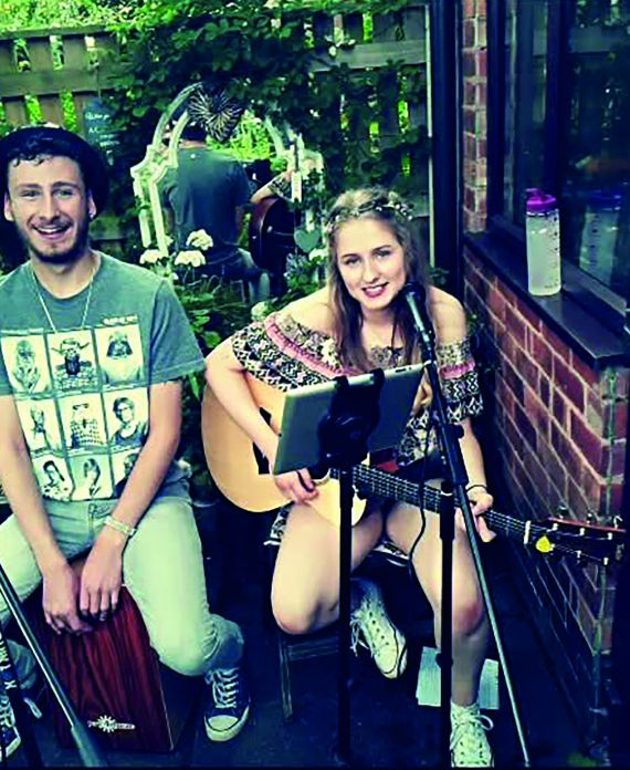 Charlie wearing a hat on the left smiling at the camera. Keria is holding a guitar and wearing a flower crown and smiling at the camera. They are both sat in the garden.