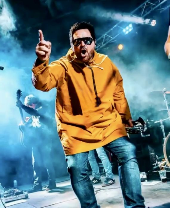 Oasis2ribute lead singer in a yellow jacket and looking at the camera with index finger pointed upwards.