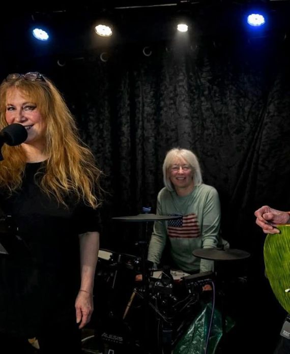 A picture of the band Let's HAve Fun playing on a stage of black curtains in the background. There are four members of the band playing.